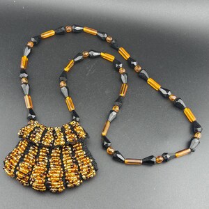 Può includere: Una collana di perline con un ciondolo nero e oro. La collana presenta un ciondolo a forma di pettorina con file di perline dorate e un retro in rete nera. La catena è composta da perline nere e color ambra.