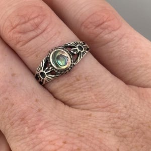 Op de afbeelding: Een zilveren ring met een centrale ovale abalone schelp inleg in iriserende groen- en blauwtinten. De band is aan beide zijden van de steen versierd met gegraveerde zonnestraalpatronen, wat zorgt voor een gedetailleerd metalen ontwerp rond de vinger.