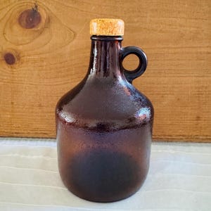 May include: A brown glass jug with a handle and a cork stopper. The jug has a rounded body and a narrow neck. The cork is light brown. The background is a wooden surface.