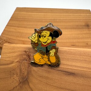 May include: A colorful enamel pin featuring Mickey Mouse in a cowboy outfit. Mickey is wearing a brown cowboy hat, a blue jacket, yellow pants, and a red bandana. He is holding a bag of gold. The pin is set against a wooden background.