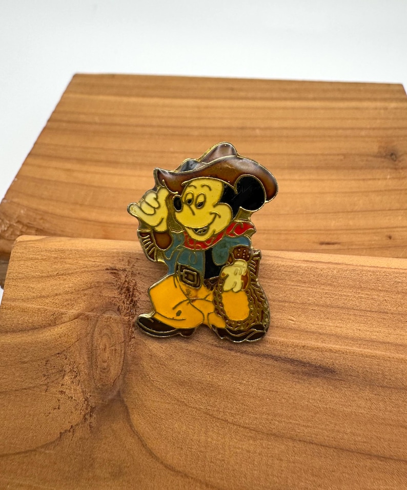 May include: A colorful enamel pin featuring Mickey Mouse in a cowboy outfit. Mickey is wearing a brown cowboy hat, a blue jacket, yellow pants, and a red bandana. He is holding a bag of gold. The pin is set against a wooden background.