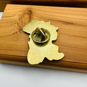 May include: A gold-colored metal pin with a map outline. The pin has a butterfly clutch on the back for secure attachment. The pin is set against a wooden surface.