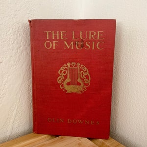 May include: A red hardcover book titled "The Lure of Music" by Olin Downes. The book cover features a gold illustration of a lyre.