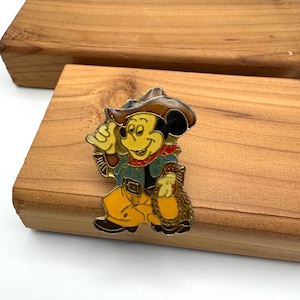 May include: A colorful enamel pin featuring Mickey Mouse dressed as a cowboy. Mickey is wearing a brown hat, blue jacket, yellow pants, and a red scarf. He is holding a bag of gold coins. The pin is set against a wooden background.