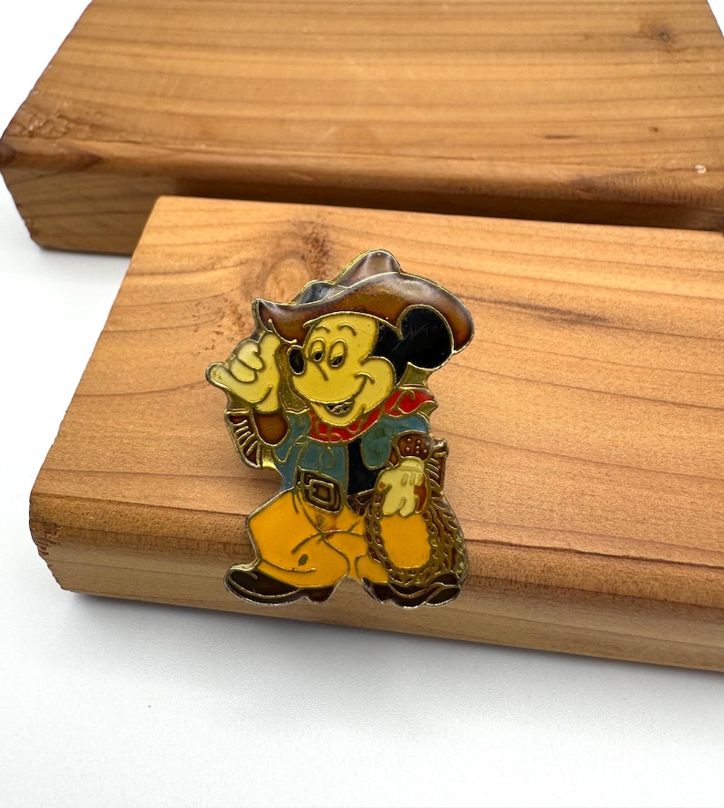 May include: A colorful enamel pin featuring Mickey Mouse dressed as a cowboy. Mickey is wearing a brown hat, blue jacket, yellow pants, and a red scarf. He is holding a bag of gold coins. The pin is set against a wooden background.
