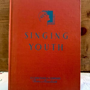 May include: A vintage book titled "Singing Youth" with a red cover and teal lettering. The cover features a teal illustration of a profile and a square. The publisher's name and location are printed at the bottom.