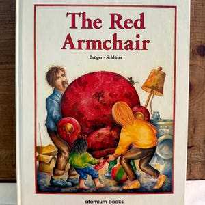 May include: A children's book cover titled "The Red Armchair" by Brüger - Schlüter. The cover features a red armchair being moved by three people. The armchair is large and appears to be a statement piece.