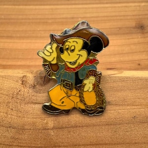 May include: A vintage enamel pin featuring Mickey Mouse dressed as a cowboy. The pin has a brown cowboy hat, a blue shirt, yellow pants, and a red bandana. Mickey is holding a rope and giving a thumbs-up.