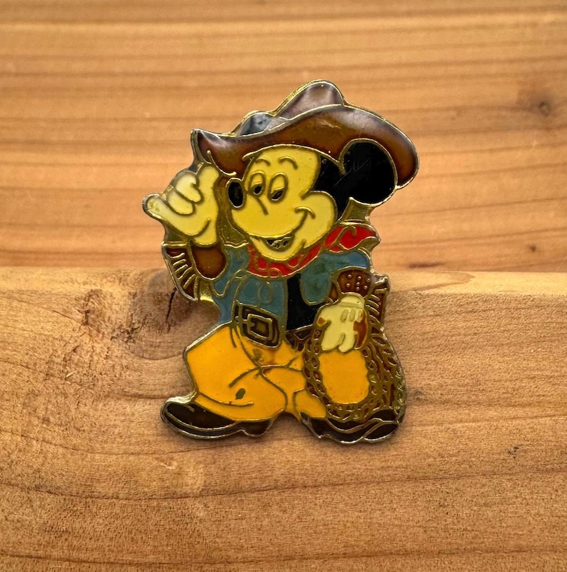 May include: A vintage enamel pin featuring Mickey Mouse dressed as a cowboy. The pin has a brown cowboy hat, a blue shirt, yellow pants, and a red bandana. Mickey is holding a rope and giving a thumbs-up.