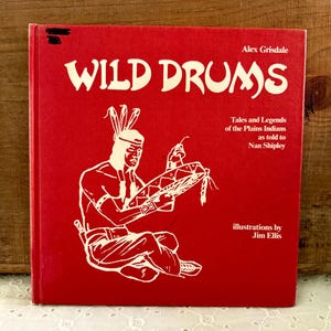 May include: A red hardcover book titled "WILD DRUMS" with white text. The book cover features an illustration of a Native American man playing a drum. The text on the cover reads "Tales and Legends of the Plains Indians as told to Nan Shipley" and "illustrations by Jim Ellis".