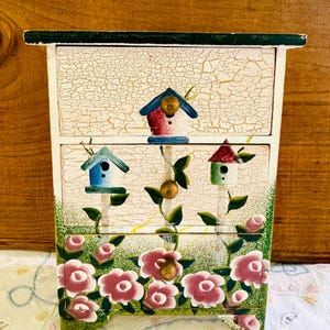 May include: A small wooden box with three drawers painted with a whimsical design of birdhouses and roses. The box has a distressed white paint finish with green accents.