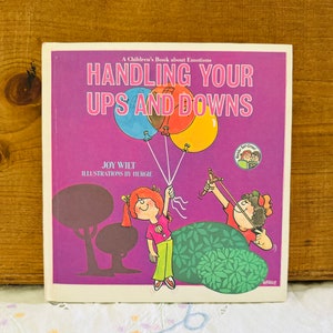 May include: A purple book cover with the title "Handling Your Ups and Downs" in white text. The book is about emotions and features illustrations of children with balloons and a slingshot.