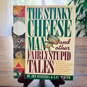 May include: A hardcover book titled "The Stinky Cheese Man and Other Fairly Stupid Tales" is displayed on a wooden stand. The book cover features a cartoon cheese character and a list of stories. The book is by Jon Scieszka & Lane Smith.