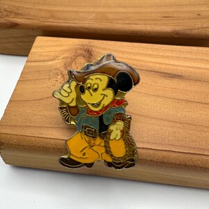 May include: A vintage enamel pin featuring Mickey Mouse in a cowboy outfit. Mickey is wearing a brown cowboy hat, blue jacket, yellow pants, and a red neckerchief. He is holding a bag of gold. The pin is set against a wooden background.