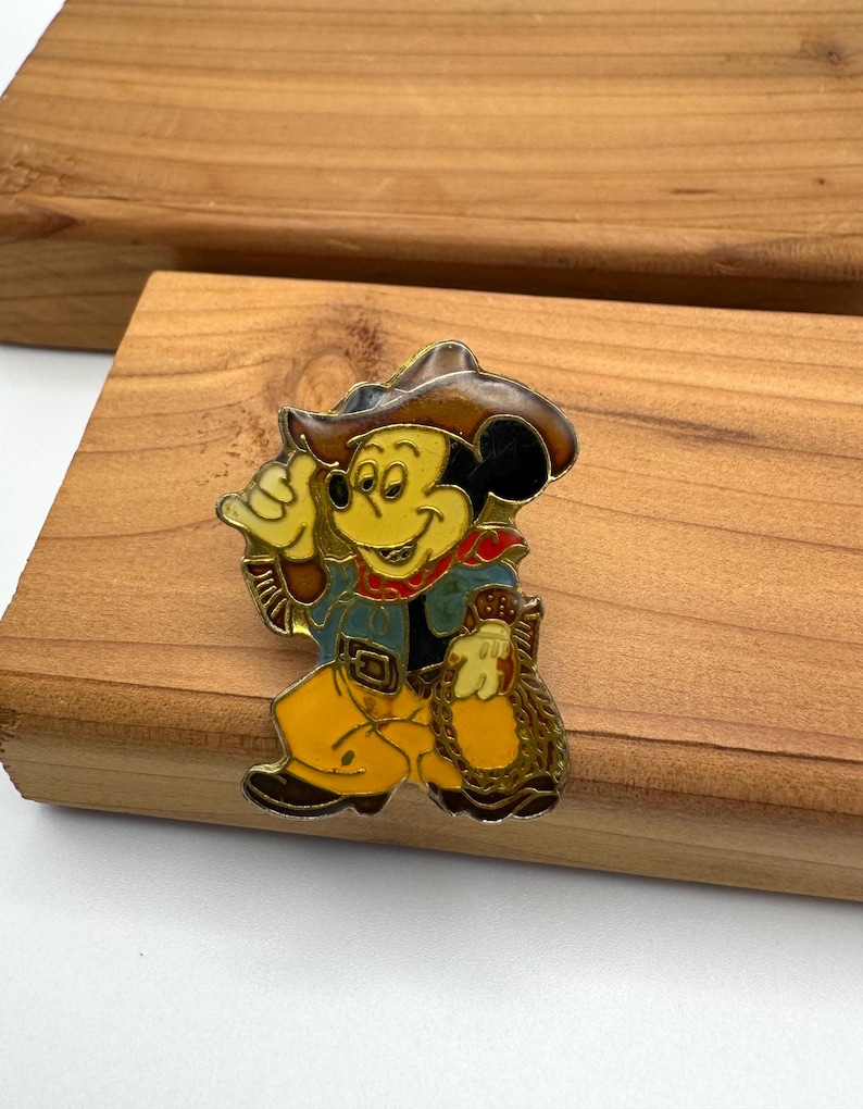 May include: A vintage enamel pin featuring Mickey Mouse in a cowboy outfit. Mickey is wearing a brown cowboy hat, blue jacket, yellow pants, and a red neckerchief. He is holding a bag of gold. The pin is set against a wooden background.