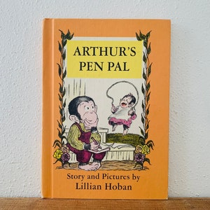 May include: A vintage children's book titled "Arthur's Pen Pal" by Lillian Hoban. The cover is orange with illustrations of a monkey writing and another jumping rope. The title is in yellow, and the book is set on a wooden surface.