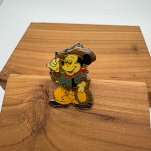 May include: A colorful enamel pin featuring Mickey Mouse dressed as a cowboy. Mickey is wearing a brown hat, blue jacket, yellow pants, and a red neckerchief. He is holding a rope and a bag.