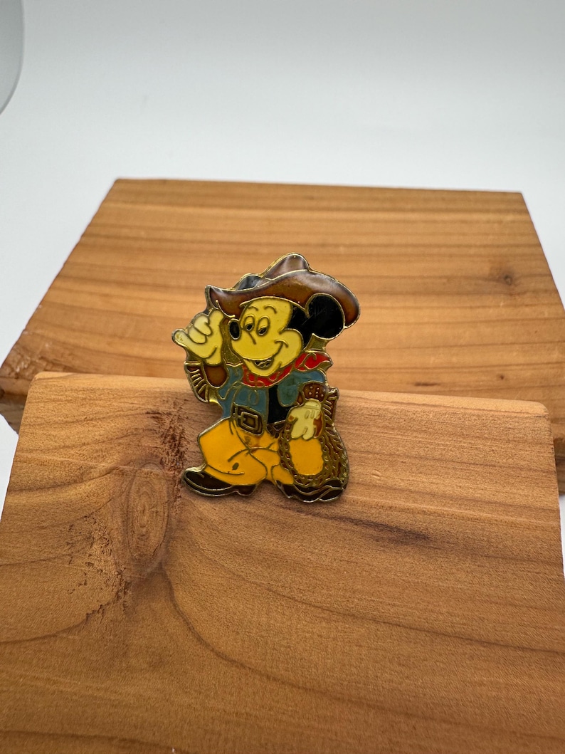 May include: A colorful enamel pin featuring Mickey Mouse dressed as a cowboy. Mickey is wearing a brown hat, blue jacket, yellow pants, and a red neckerchief. He is holding a rope and a bag.