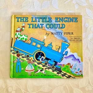 May include: A vintage children's book cover featuring a blue train engine with a smiling face, pulling a train car up a hill. The book title is "The Little Engine That Could" by Watty Piper. The cover is yellow with green and purple accents.