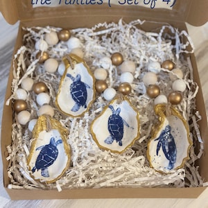 May include: A set of four oyster shell napkin rings, each featuring a blue turtle design on a white background with gold trim. The shells are displayed in a cardboard box with decorative beads and paper filler. The text "Napkin Rings...All about the Turtles (Set of 4)" is visible.