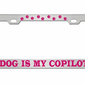 May include: A silver licence plate frame with pink paw prints along the top and the text "DOG IS MY COPILOT" in pink letters at the bottom.