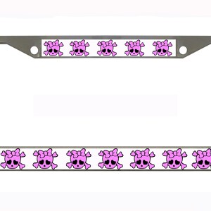 Puede incluir: Un marco de matrícula plateado con un borde blanco que presenta un patrón repetido de calaveras rosas con lazos. Las calaveras tienen ojos negros sobre un fondo blanco.