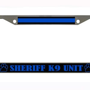 Puede incluir: Un marco de matrícula plateado con una franja negra y azul en la parte superior y las palabras "SHERIFF K9 UNIT" en azul. Huellas de patas flanquean el texto.