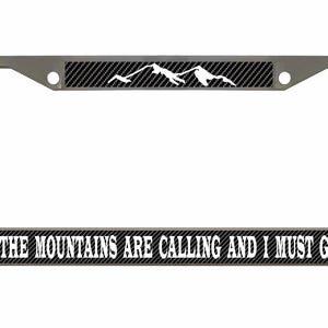 May include: A silver license plate frame with a black carbon fiber background. The top of the frame has a white mountain silhouette. The bottom of the frame has the text "THE MOUNTAINS ARE CALLING AND I MUST GO" in white.