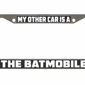 Puede incluir: Un marco de matrícula de coche plateado con un fondo de fibra de carbono negro. El texto "MY OTHER CAR IS A THE BATMOBILE" está impreso en blanco en el marco.