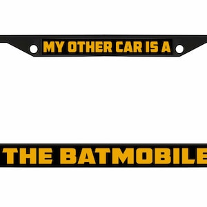 Puede incluir: Marco negro para matrícula con el texto "MY OTHER CAR IS A" en la parte superior y "THE BATMOBILE" en la parte inferior, en letras amarillas. El marco es rectangular.