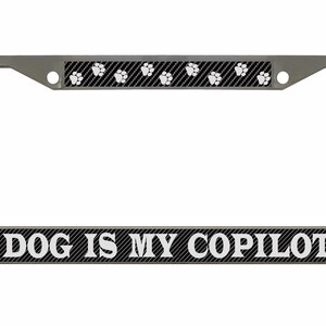 May include: A silver metal licence plate frame with a black carbon fibre pattern and white paw prints. The bottom of the frame has the text "DOG IS MY COPILOT" in white letters.