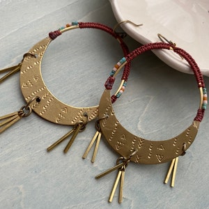 May include: A pair of gold hoop earrings with a crescent moon design and beaded accents. The earrings have a burgundy cord and multiple gold tassels.