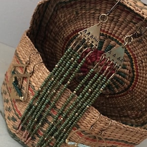 May include: A pair of long, green beaded earrings with gold accents and triangle-shaped tops. The earrings are displayed in a woven basket.