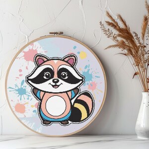 Peut inclure: Un motif de point de croix représentant un raton laveur de dessin animé avec un corps rose, des rayures noires et blanches et une chemise bleue. Le raton laveur est entouré d'éclaboussures de peinture colorées sur un fond violet clair.