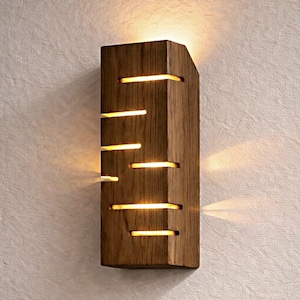 A vertical rectangular wooden wall sconce with warm LED light shining through carved horizontal openings. The wood has a natural grain and rustic brown finish. The style is minimalist, cozy, and modern, suitable for hallway, living room, or bedroom wall lighting.