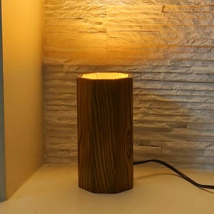 Puede incluir: Una lámpara de mesa de madera que emite una luz cálida. La lámpara tiene forma cilíndrica con un patrón de vetas de madera texturizadas. La lámpara está conectada a una toma de corriente con un cable negro. El fondo presenta una pared de piedra texturizada.