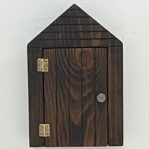Puede incluir: Un pequeño porta llaves de madera con forma de casa, con un acabado marrón oscuro. La puerta tiene dos bisagras de latón y un pomo plateado.