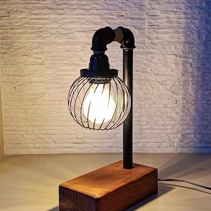 Puede incluir: Lámpara de mesa de estilo industrial con base de madera y detalles de tubería metálica negra. La lámpara presenta una pantalla de jaula de alambre que rodea una bombilla brillante. La base es un bloque rectangular de madera teñida.