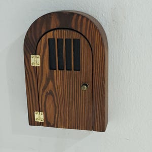 Può includere: Una porta delle fate in legno con la parte superiore ad arco, con una piccola porta con pomello e cerniere in ottone. La porta ha una macchia scura e doghe nere verticali sopra la porta. La porta è montata su un muro bianco.