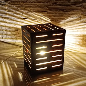 May include: A wooden lamp with a geometric design. The lamp is lit and casts a warm glow. The lamp is sitting on a tiled surface.