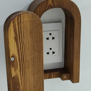 May include: Wooden electrical outlet cover with a hinged door. The cover is made of stained wood and has a rounded arch shape. The white electrical outlet has three plug receptacles.