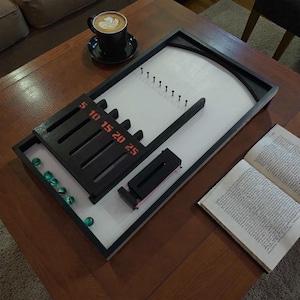 Peut inclure: Un jeu de table avec un cadre noir, une surface de jeu blanche et des éléments de score noirs. Le jeu comprend une série de broches, une rampe et un système de pointage marqué de chiffres rouges. Plusieurs billes vertes sont visibles. Une tasse de café et un livre sont également sur la table.