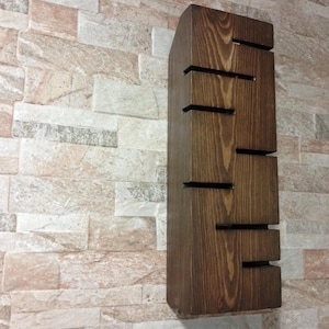 A handmade wooden wall lamp with warm ambient lighting. The lamp has vertical rectangular shape with carved horizontal openings that allow light to shine outward. The wood has a natural rustic texture and dark brown finish. The design is minimalist, modern, and Scandinavian-inspired, perfect for cozy home decor or hallway lighting.