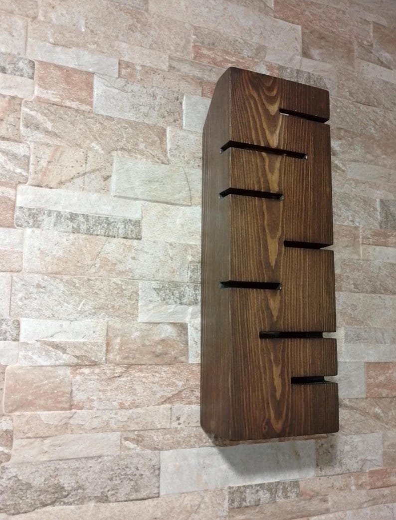 A handmade wooden wall lamp with warm ambient lighting. The lamp has vertical rectangular shape with carved horizontal openings that allow light to shine outward. The wood has a natural rustic texture and dark brown finish. The design is minimalist, modern, and Scandinavian-inspired, perfect for cozy home decor or hallway lighting.