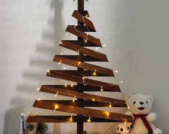May include: A wooden Christmas tree with a gold star topper and string lights. The tree is decorated with red ornaments and sits on a white, fringed table runner. A white teddy bear with a red scarf is next to the tree.