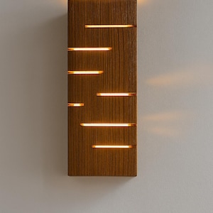 A vertical rectangular wooden wall sconce with warm LED light shining through carved horizontal openings. The wood has a natural grain and rustic brown finish. The style is minimalist, cozy, and modern, suitable for hallway, living room, or bedroom wall lighting.