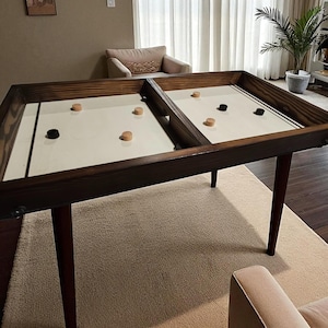 Puede incluir: Una mesa de shuffleboard de madera con marco y patas de color marrón oscuro. La superficie de juego es blanquecina con discos de madera y negros. La mesa está sobre una alfombra beige en una habitación bien iluminada.
