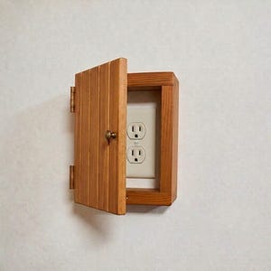 Puede incluir: Una cubierta rectangular de madera con una puerta abatible que se abre para revelar un enchufe de pared blanco estándar. La cubierta está construida con tablones de madera verticales y cuenta con un pequeño pomo redondo y dos bisagras metálicas.