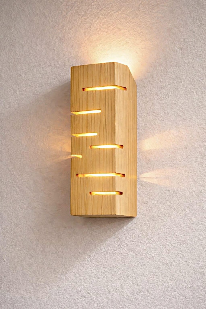 A vertical rectangular wooden wall sconce with warm LED light shining through carved horizontal openings. The wood has a natural grain and rustic brown finish. The style is minimalist, cozy, and modern, suitable for hallway, living room, or bedroom wall lighting.