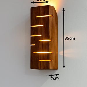 A vertical rectangular wooden wall sconce with warm LED light shining through carved horizontal openings. The wood has a natural grain and rustic brown finish. The style is minimalist, cozy, and modern, suitable for hallway, living room, or bedroom wall lighting.
